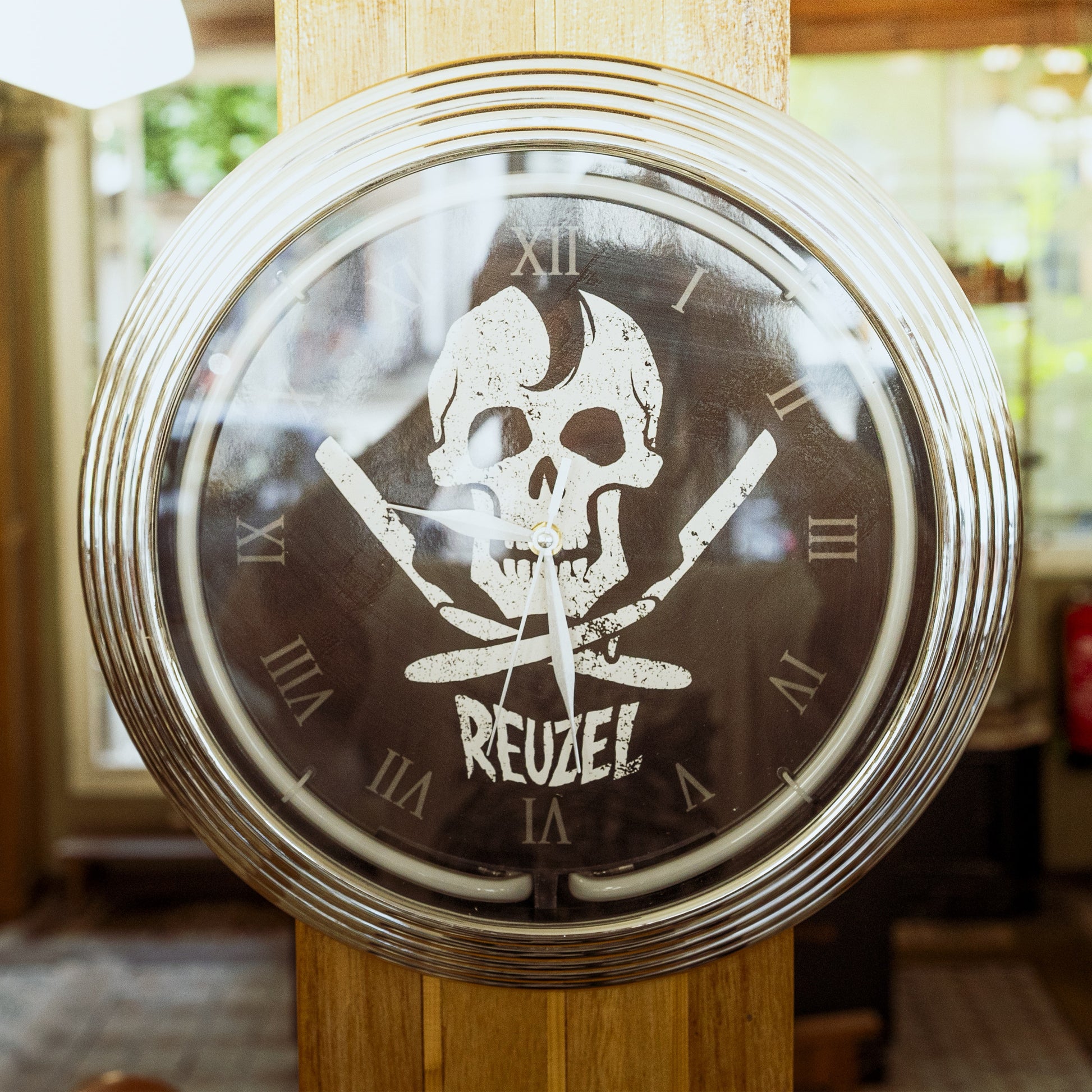 A round wall clock with a metallic silver frame and a dark face featuring a white skull and crossed knives above the word 'REUZEL'. The clock is mounted on a wooden post in a real indoor setting with blurred background details.